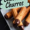 Close-up of chocolate-dipped churros arranged on parchment paper over a dark marble surface. The churros have a golden-brown crispy texture with glossy chocolate coating the ends. A text overlay reads "Chocolate Churros" in bold lettering on a mint green background.