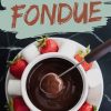 Overhead view of a bowl filled with rich, melted chocolate fondue surrounded by a variety of dippers, including vibrant red strawberries and fluffy white marshmallows, all arranged on a white plate. A fondue fork dips a piece of fruit into the smooth, glossy chocolate. Text overlay reads “Chocolate Fondue.”