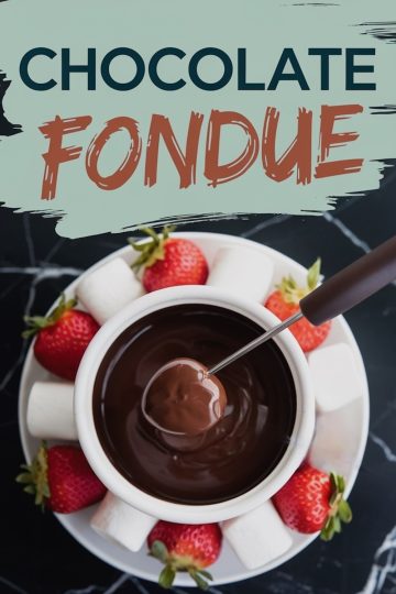 Overhead view of a bowl filled with rich, melted chocolate fondue surrounded by a variety of dippers, including vibrant red strawberries and fluffy white marshmallows, all arranged on a white plate. A fondue fork dips a piece of fruit into the smooth, glossy chocolate. Text overlay reads “Chocolate Fondue.”
