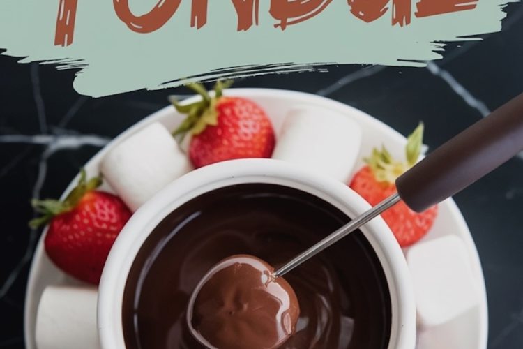 Overhead view of a bowl filled with rich, melted chocolate fondue surrounded by a variety of dippers, including vibrant red strawberries and fluffy white marshmallows, all arranged on a white plate. A fondue fork dips a piece of fruit into the smooth, glossy chocolate. Text overlay reads “Chocolate Fondue.”