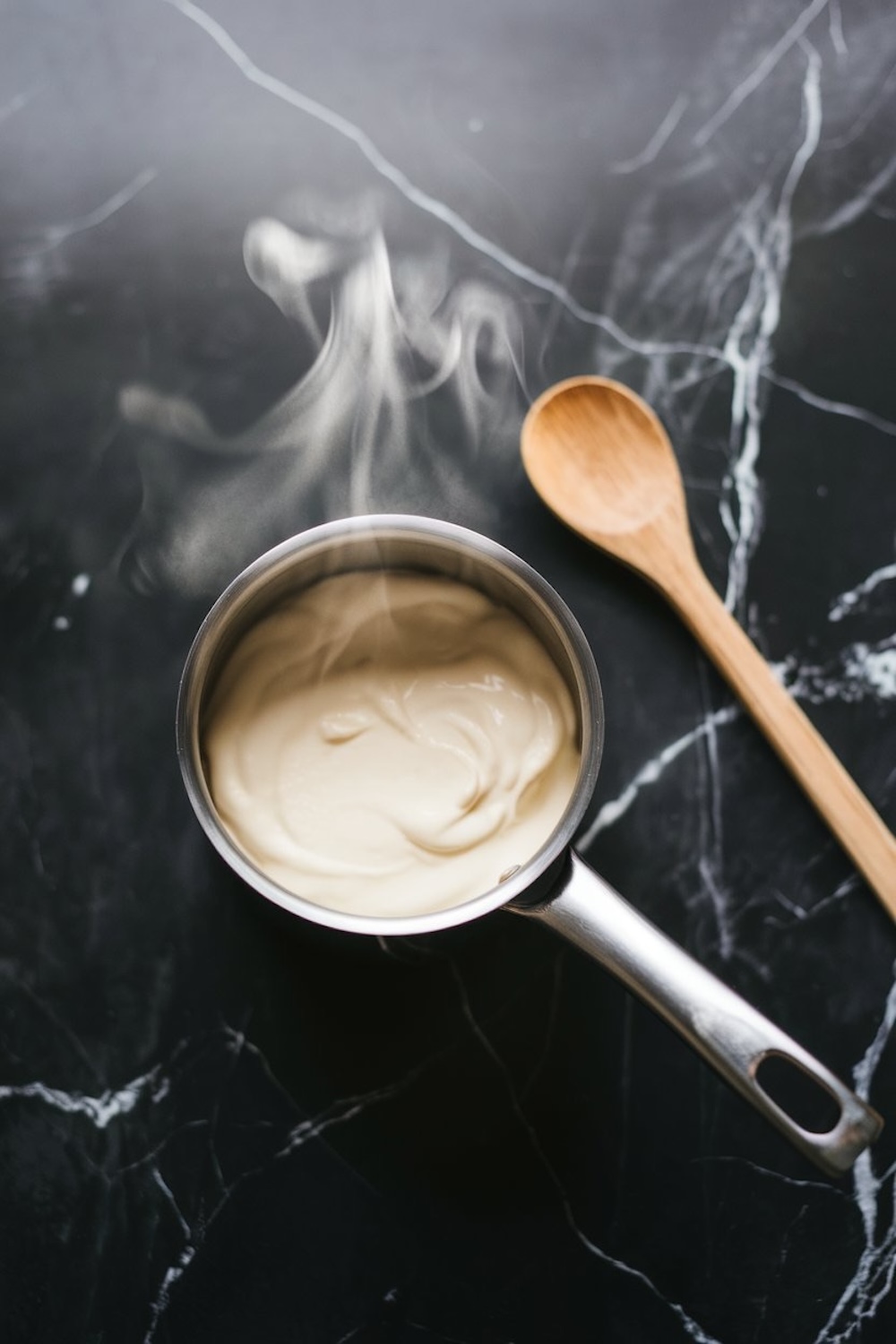  Steamy saucepan with cream being gently heated, creating visible wisps of steam rising above the pot. A wooden spoon is placed beside the saucepan, emphasizing the simplicity and freshness of the cooking process, all set on a dark marble countertop.
