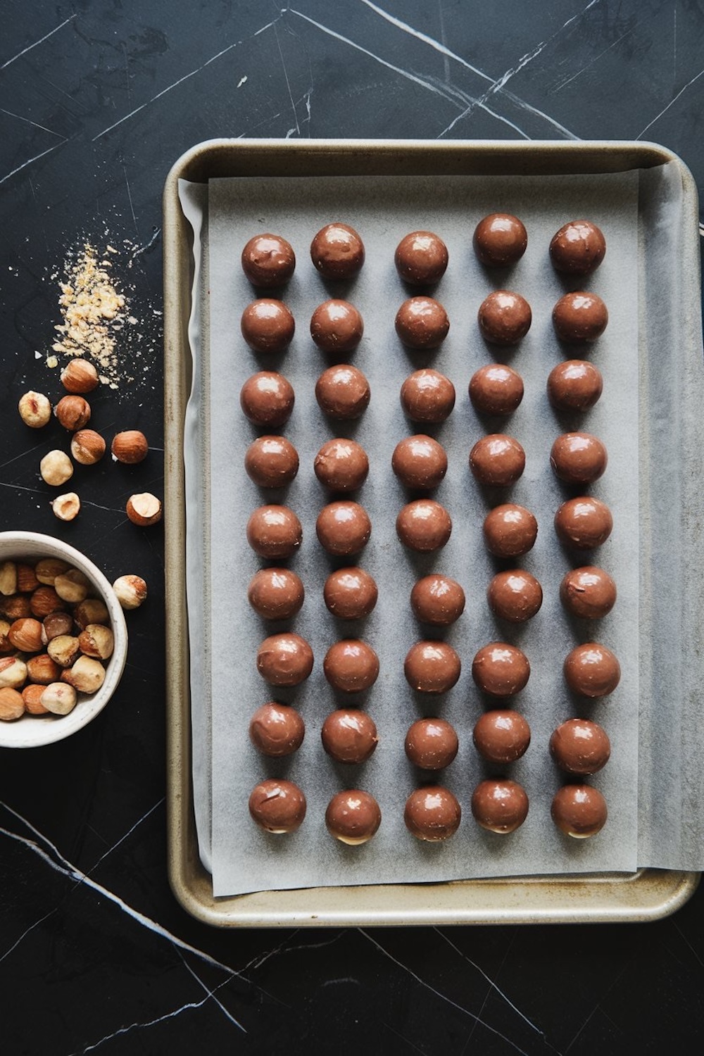 A baking tray with rows of smooth chocolate-coated truffles on parchment paper, surrounded by crushed and whole hazelnuts on a dark marble surface.