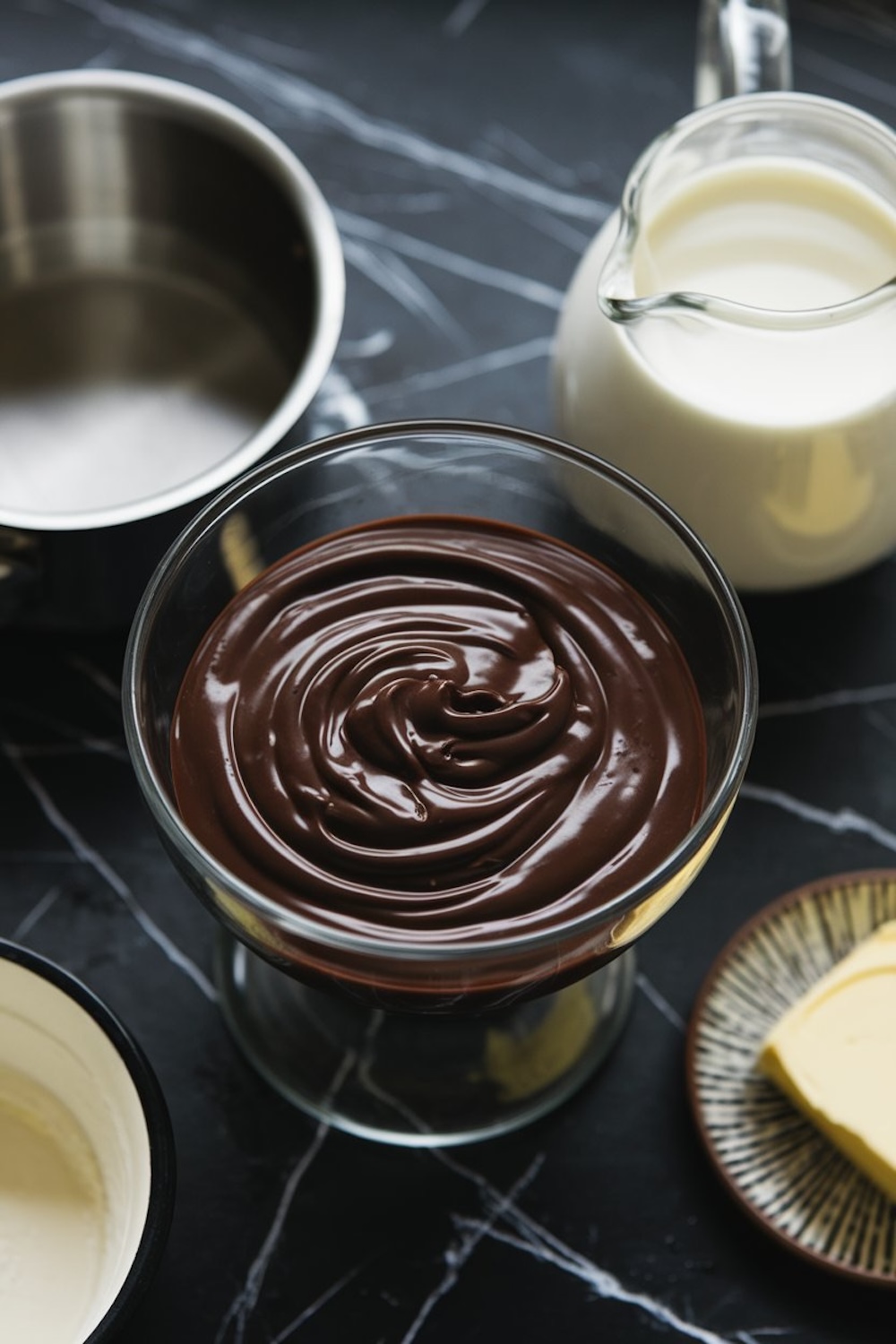 Glass bowl filled with glossy melted chocolate, surrounded by ingredients: a pitcher of milk, a dish of butter, and a saucepan, all on a dark marble countertop.