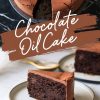A slice of rich chocolate oil cake with a tender, crackled top served on a ceramic plate. The background features the whole cake with one slice removed. Text overlay reads “Chocolate Oil Cake.”