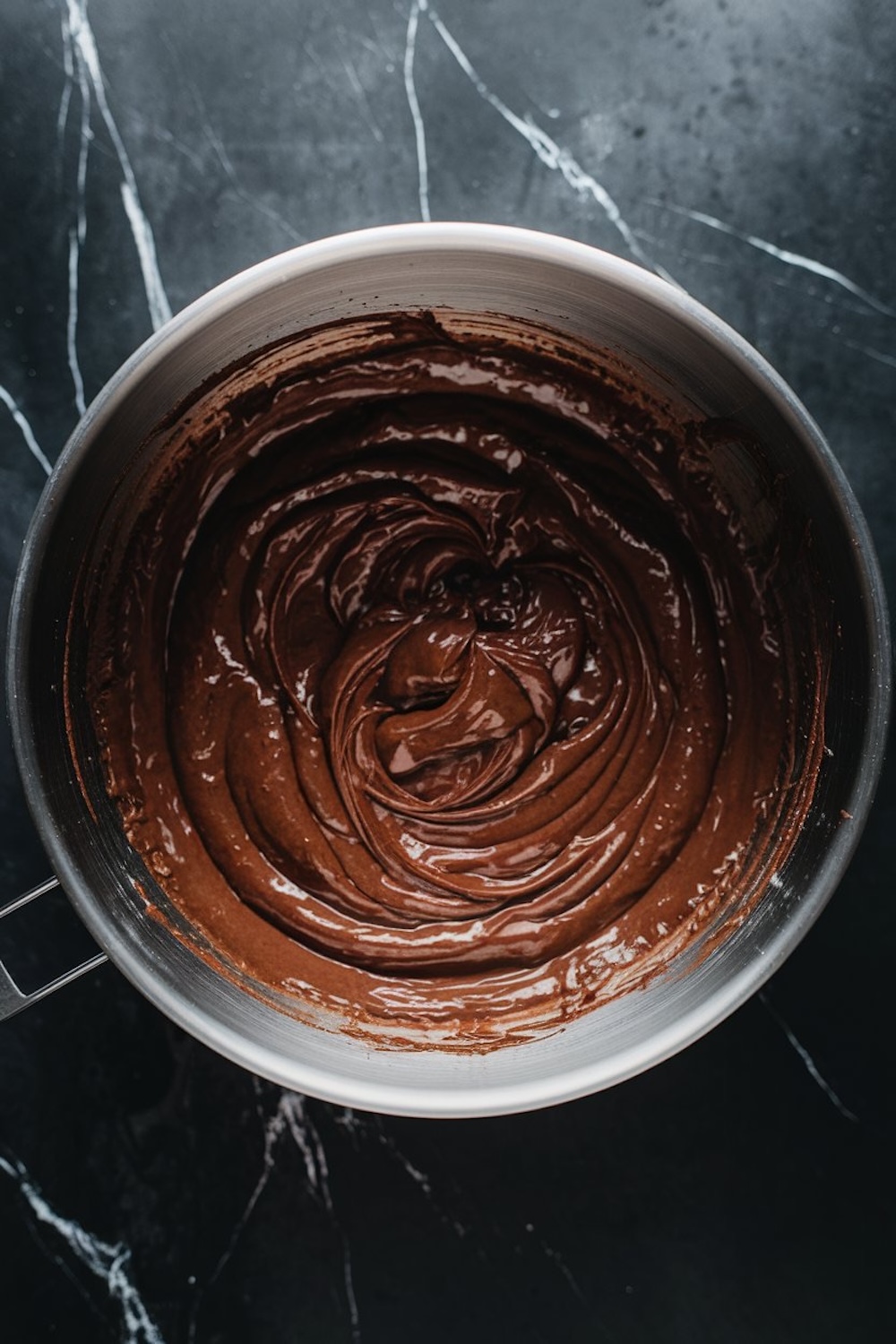 Mixing bowl filled with rich, glossy chocolate batter, capturing the creamy and thick texture against a dark marble background.
