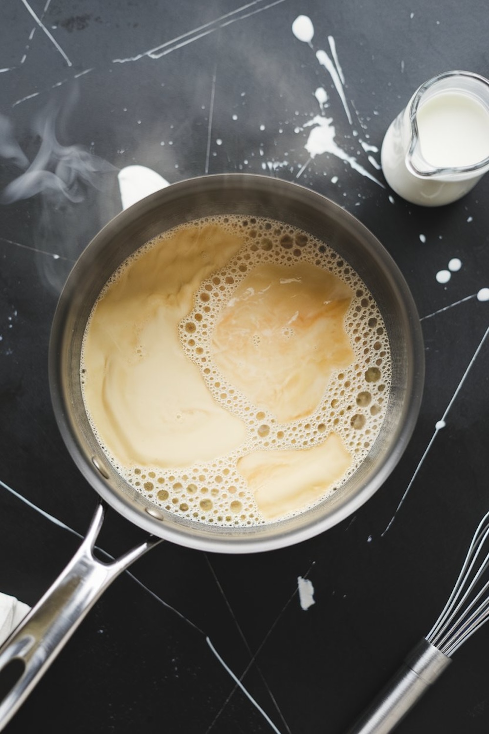 A saucepan on a black marble countertop containing simmering custard with light steam rising, surrounded by a whisk and a small pitcher of milk, showcasing the cooking process for chocolate pot de crème.