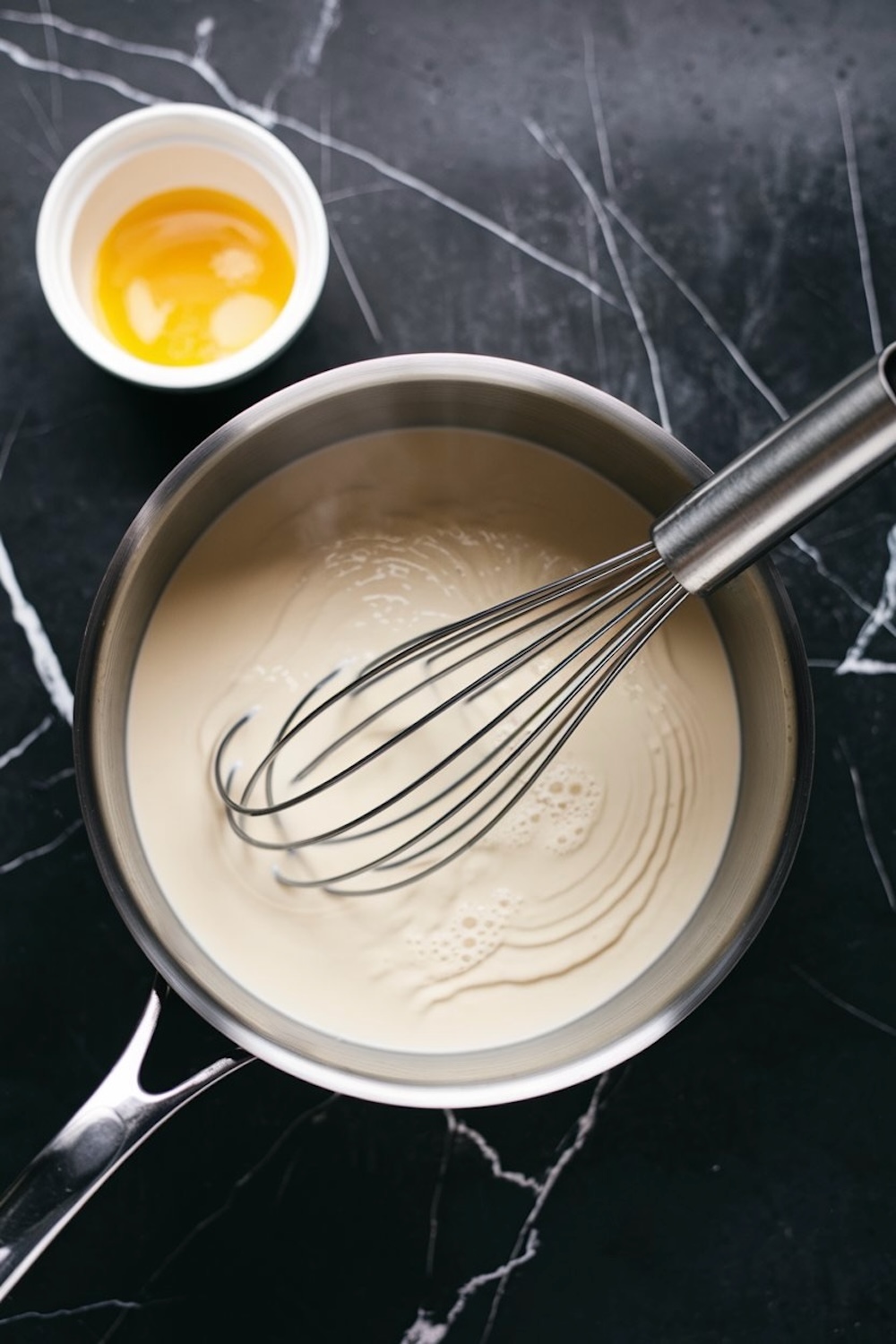 Saucepan with a whisk stirring creamy milk mixture, with a bowl of separated egg yolks in the background, set against a dark marble surface.