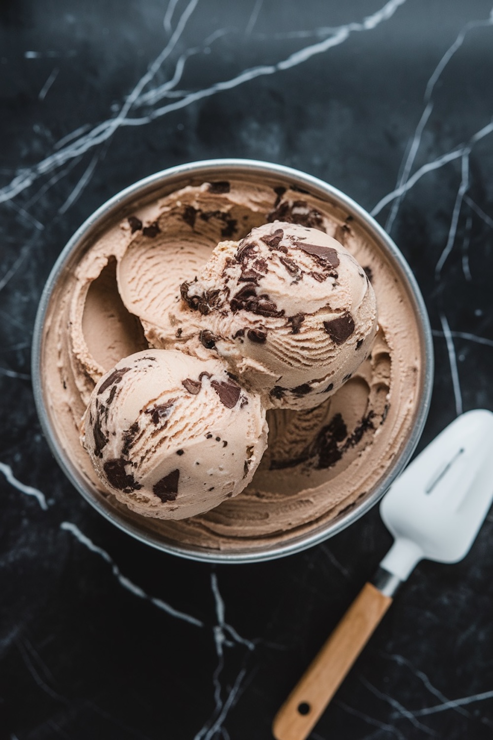 A stainless steel bowl with scoops of freshly churned coffee cookies and cream ice cream. The ice cream is rich in texture, with visible chocolate chunks, set against a dark marble background and a white ice cream scoop nearby.