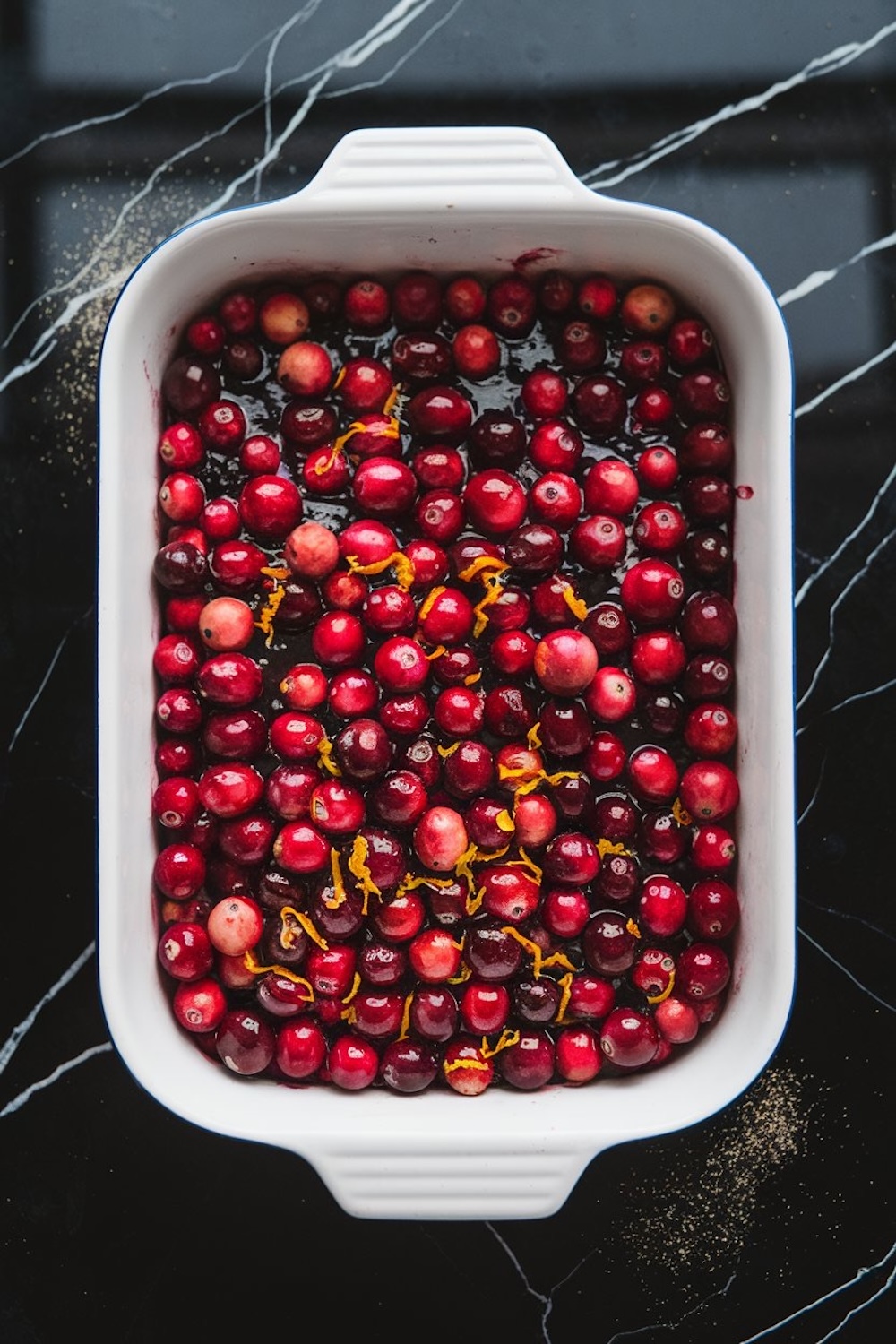 Fresh cranberries and orange zest layered in a white baking dish, ready for baking. The red cranberries and orange zest stand out against the glossy black marble countertop.