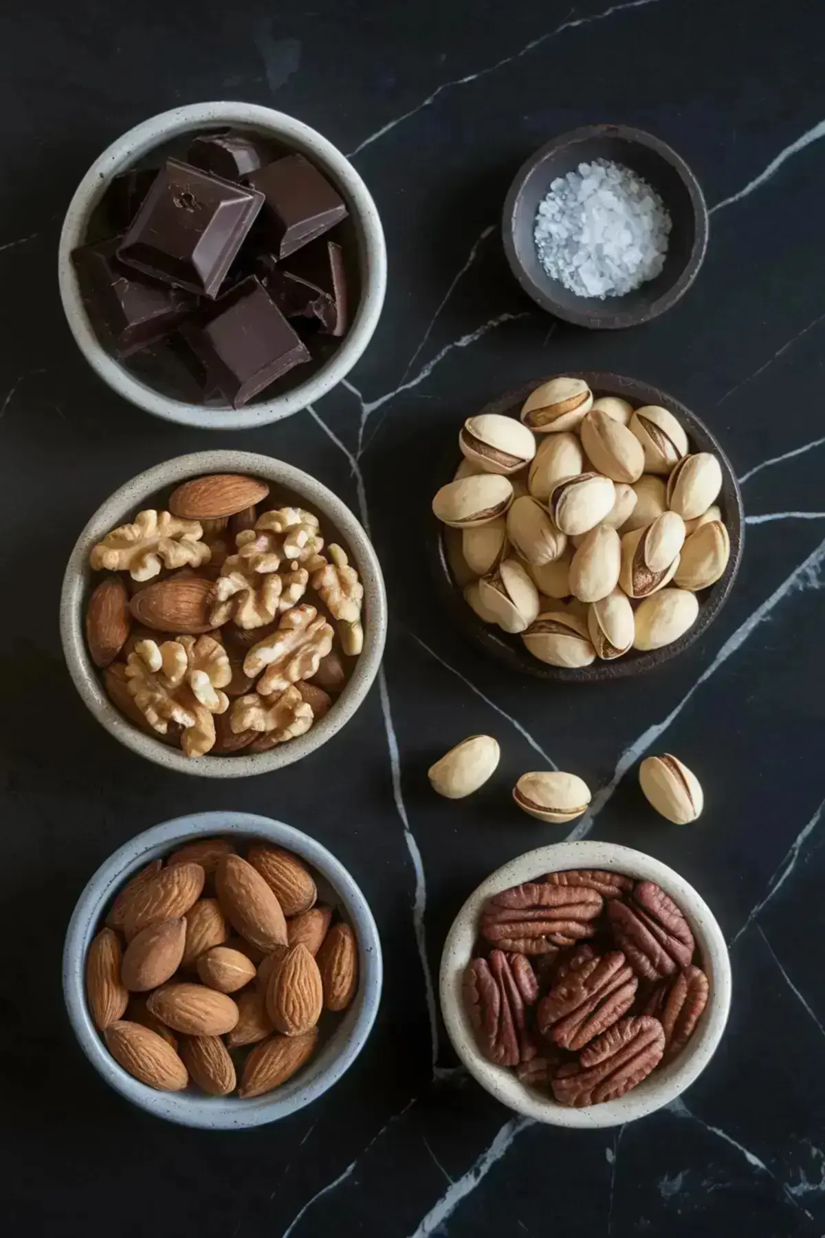 A flat lay of ingredients for dark chocolate bark, including dark chocolate chunks, coarse sea salt, and assorted nuts like almonds, walnuts, pecans, and pistachios, arranged in small bowls on a dark surface.