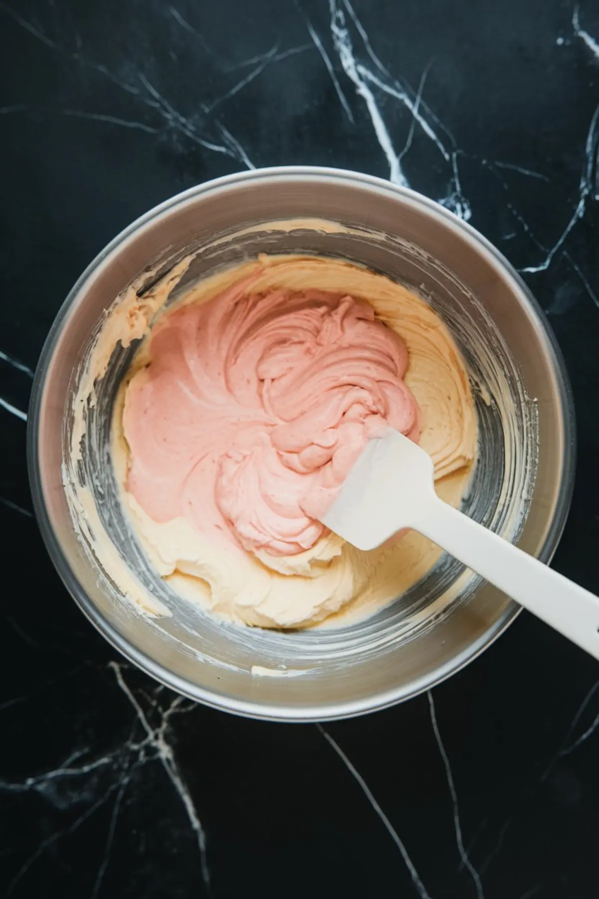 A mixing bowl with a spatula stirring the dip as pink food coloring is blended into the creamy base, creating a pastel swirl.