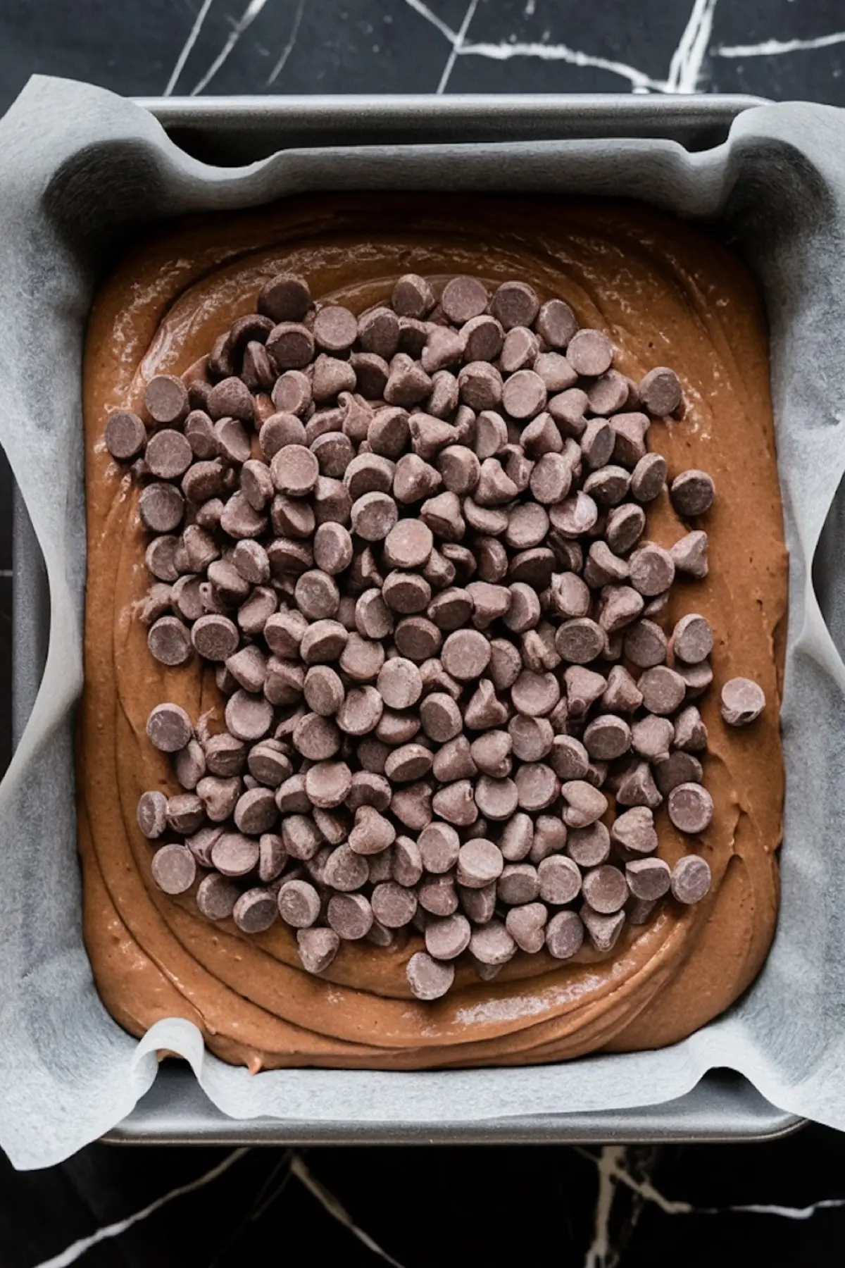 A square baking pan lined with parchment paper, filled with chocolate brownie batter topped generously with chocolate chips, ready for baking.