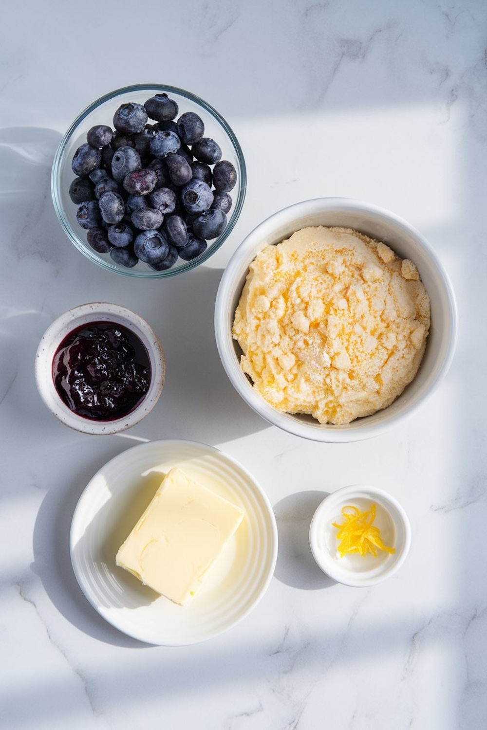 Top-down view of the ingredients for a lemon blueberry dump cake arranged on a white marble surface. The items include a bowl of fresh blueberries, a bowl of dry cake mix, a small bowl of blueberry jam, a plate with a stick of butter, and a small dish with fresh lemon zest. Natural light highlights the vibrant colors and textures.