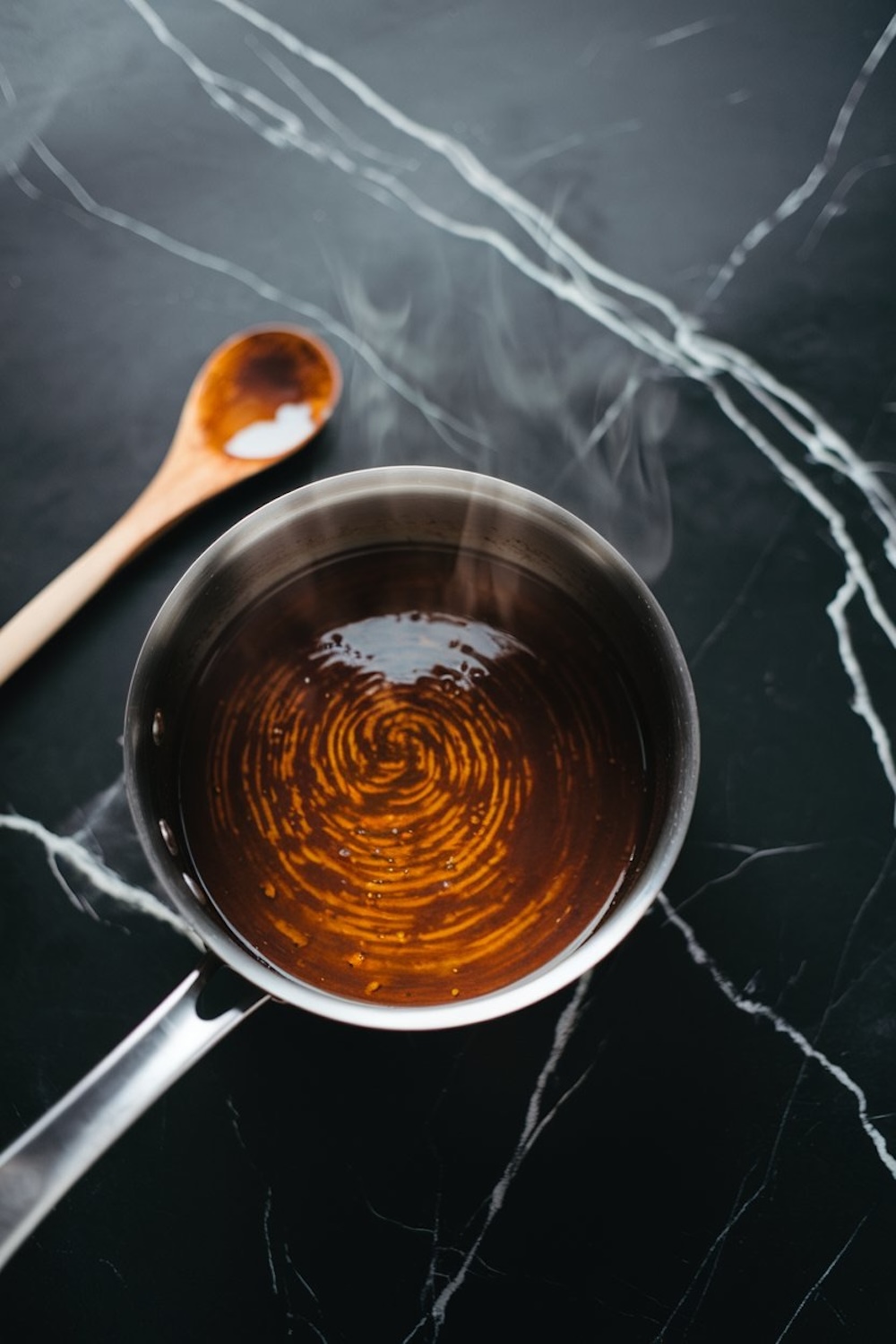 A saucepan with steaming molasses mixture, captured on a dark marble surface with a wooden spoon placed beside it.