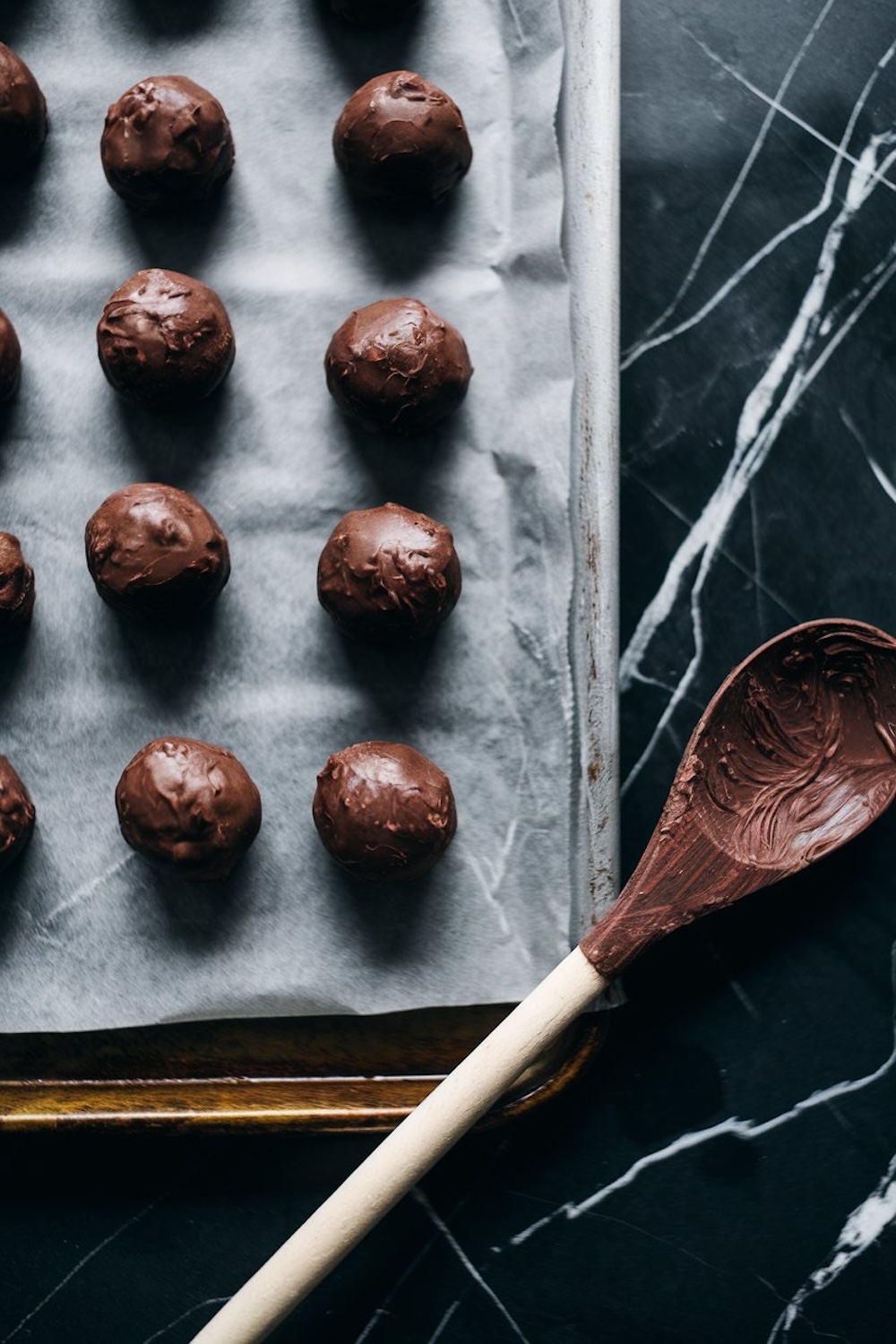 A baking tray lined with parchment paper holding rows of chocolate-coated truffle balls. A wooden spoon with chocolate residue lies beside the tray, adding a rustic preparation vibe.