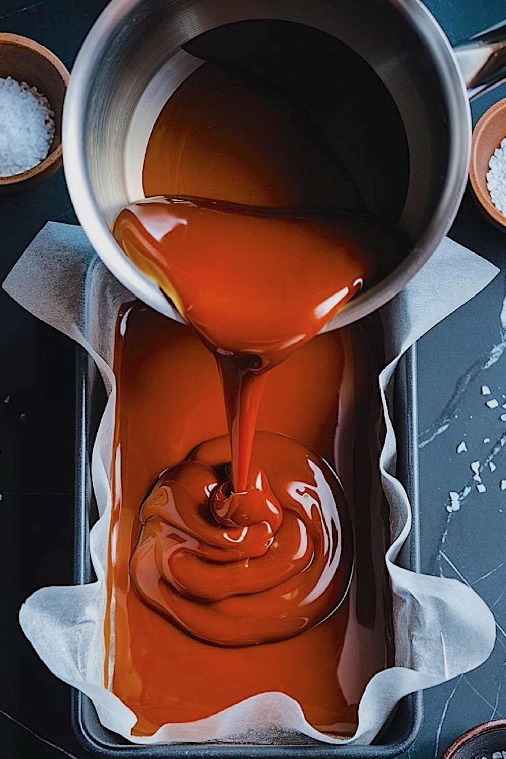 A rich caramel mixture being poured from a saucepan into a parchment-lined loaf pan, with bowls of sea salt nearby.