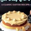 A golden-brown Simnel cake with a smooth marzipan topping, decorated with eleven toasted marzipan balls, sits on a white cake stand. A yellow ribbon is tied around the base. A hand uses a spoon to adjust one of the marzipan balls. The text overlay reads, “Simnel Cake - A Classic Easter Recipe.”