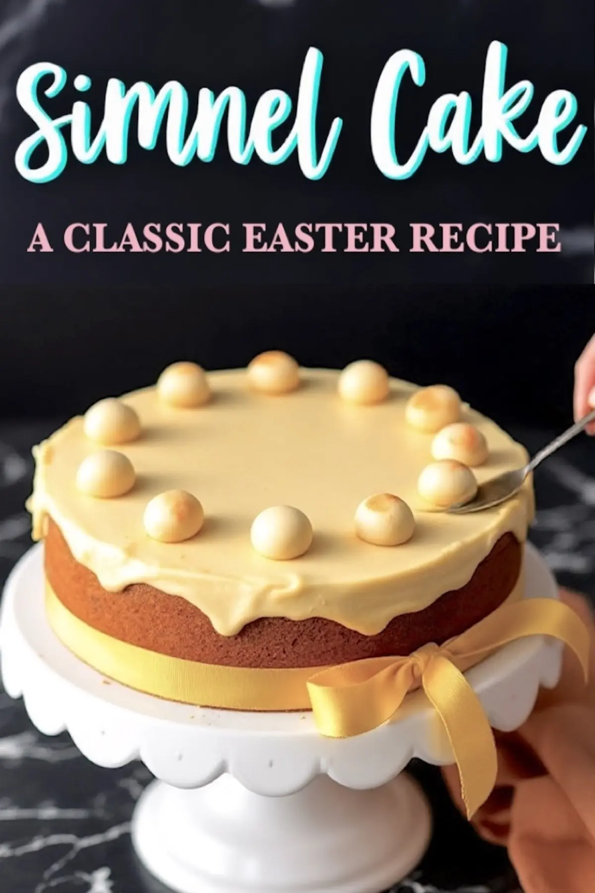 A golden-brown Simnel cake with a smooth marzipan topping, decorated with eleven toasted marzipan balls, sits on a white cake stand. A yellow ribbon is tied around the base. A hand uses a spoon to adjust one of the marzipan balls. The text overlay reads, “Simnel Cake - A Classic Easter Recipe.”