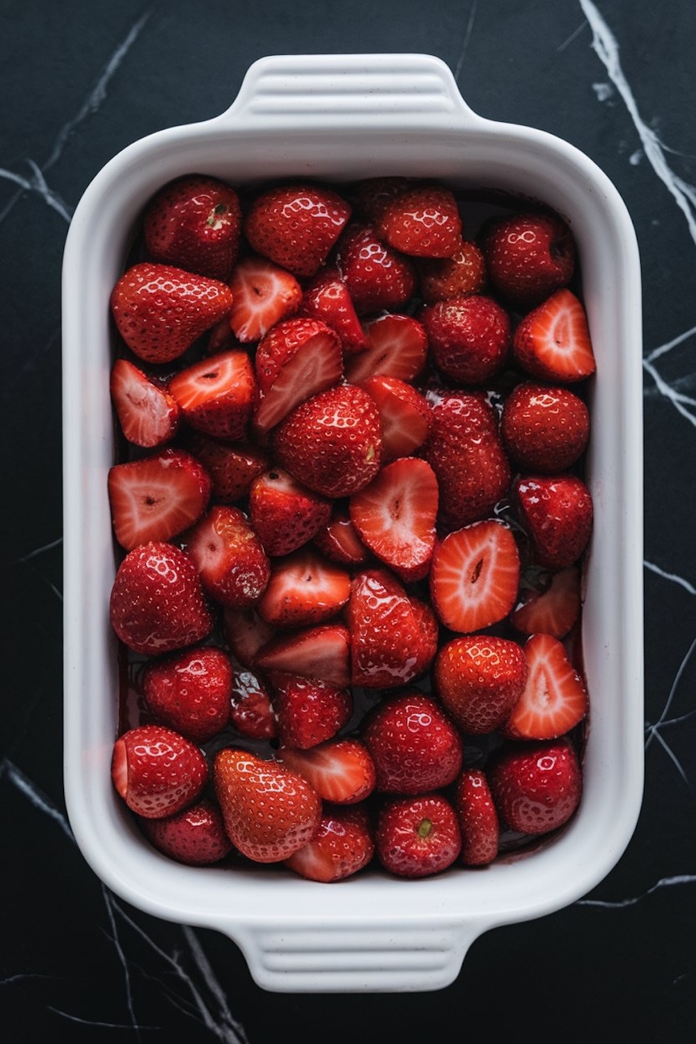A white baking dish filled with fresh halved strawberries, ready to be used as the base for a strawberry dump cake. The vibrant red strawberries contrast with the dark surface beneath.