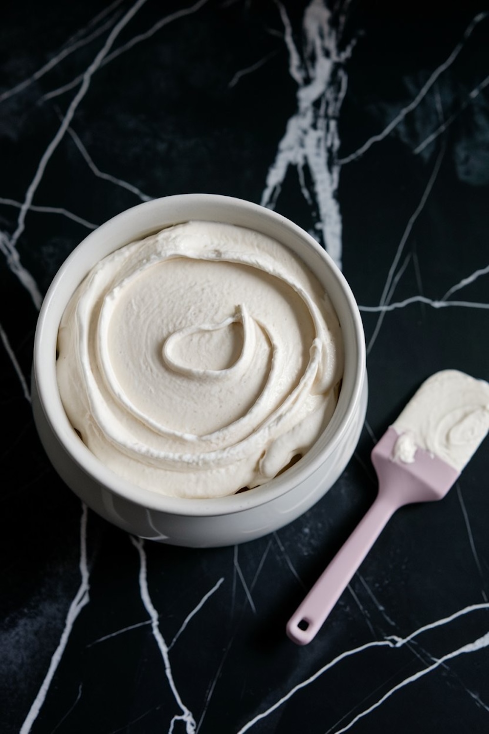 A bowl of smooth, creamy whipped filling for strawberry pretzel salad, displayed on a black marble surface with a pastel pink spatula resting beside the bowl.