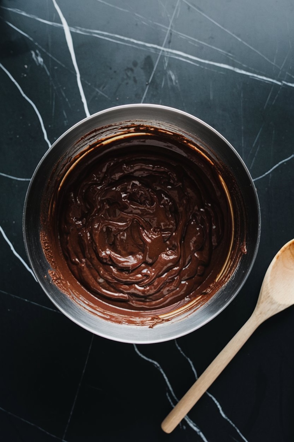 A mixing bowl filled with rich chocolate batter, placed on a black marble surface with a wooden spoon beside it.