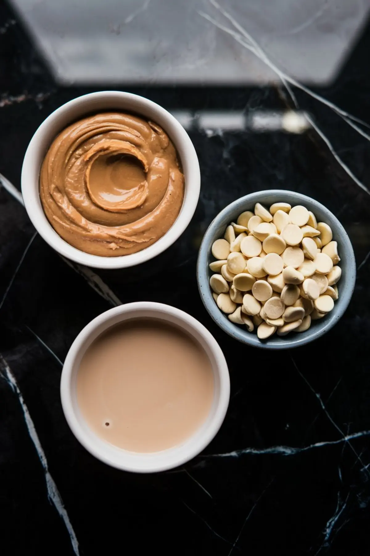 Three small bowls with ingredients for peanut butter fudge: creamy peanut butter, white chocolate chips, and a bowl of condensed milk, all arranged on a dark marble surface.