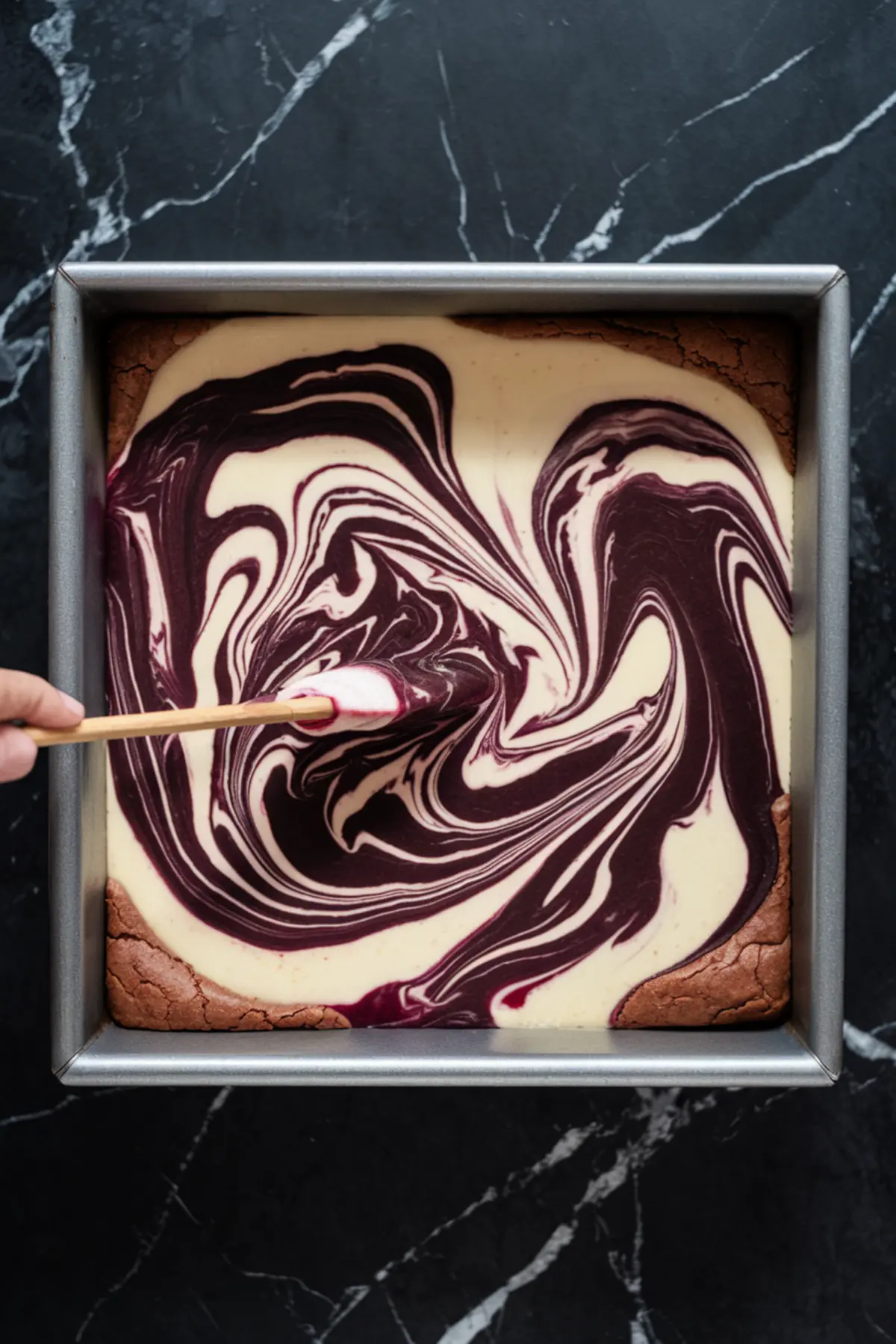 A baking pan filled with unbaked blackberry cheesecake brownie batter. A hand is using a skewer to create a marbled swirl effect, blending the blackberry sauce into the cheesecake layer.