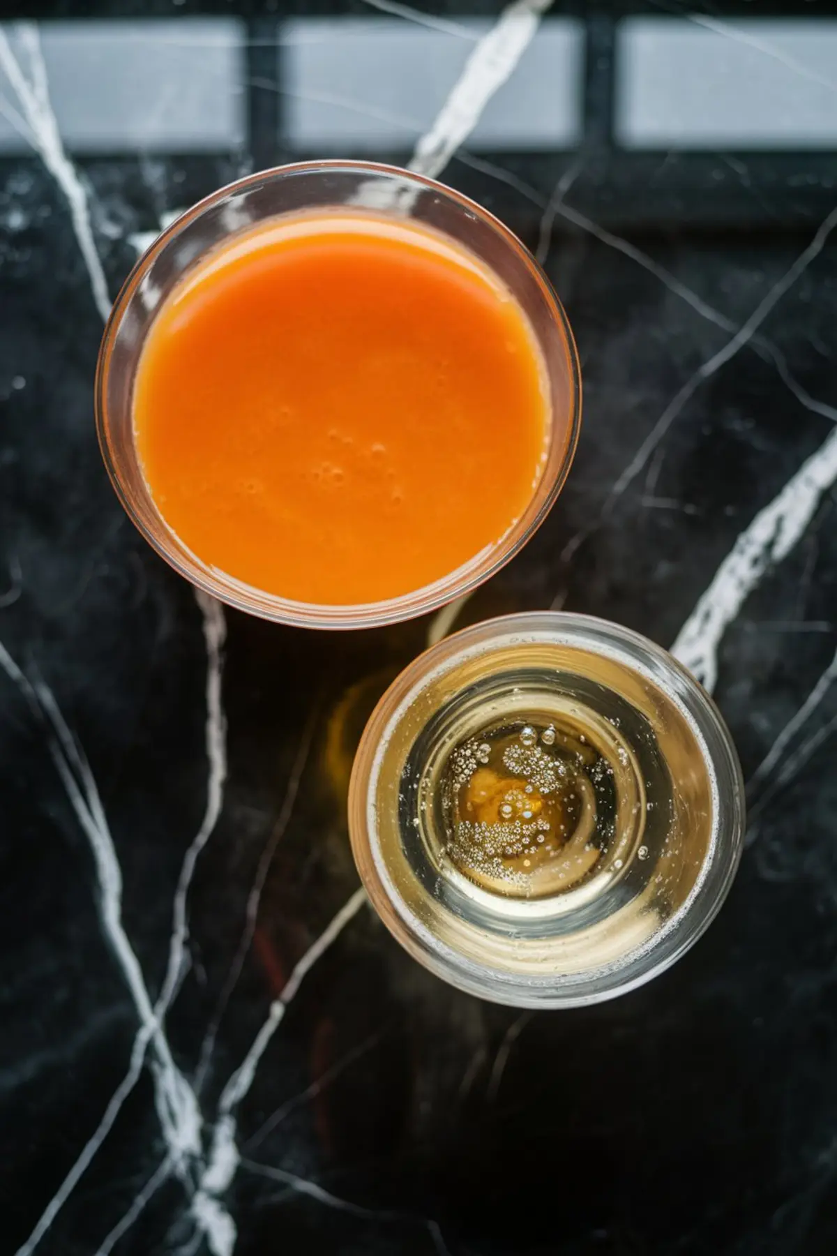 A top-down view of two glasses on a black marble countertop, one filled with orange carrot juice and the other with sparkling champagne. The contrasting colors and bubbling texture emphasize the refreshing combination used to make carrot mimosas.