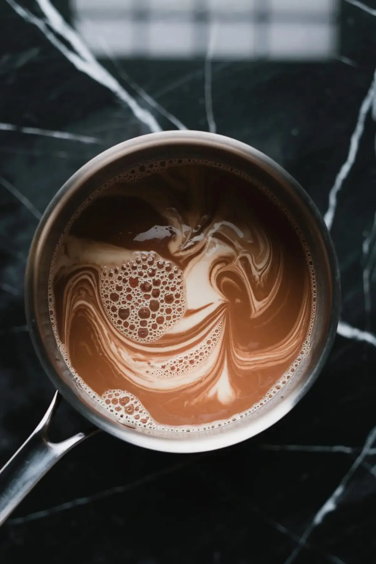 A saucepan with a swirling mixture of milk and cocoa on a black marble surface, creating a marbled effect with bubbles on the surface.