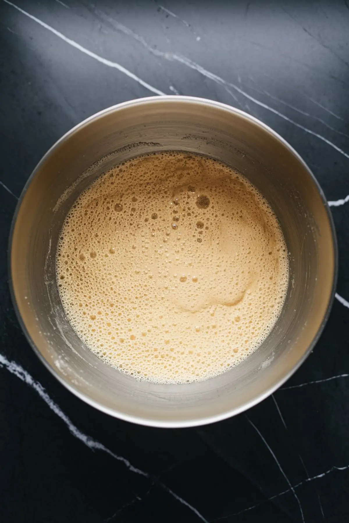 A mixing bowl filled with a frothy, light brown dough mixture, sitting on a black countertop.