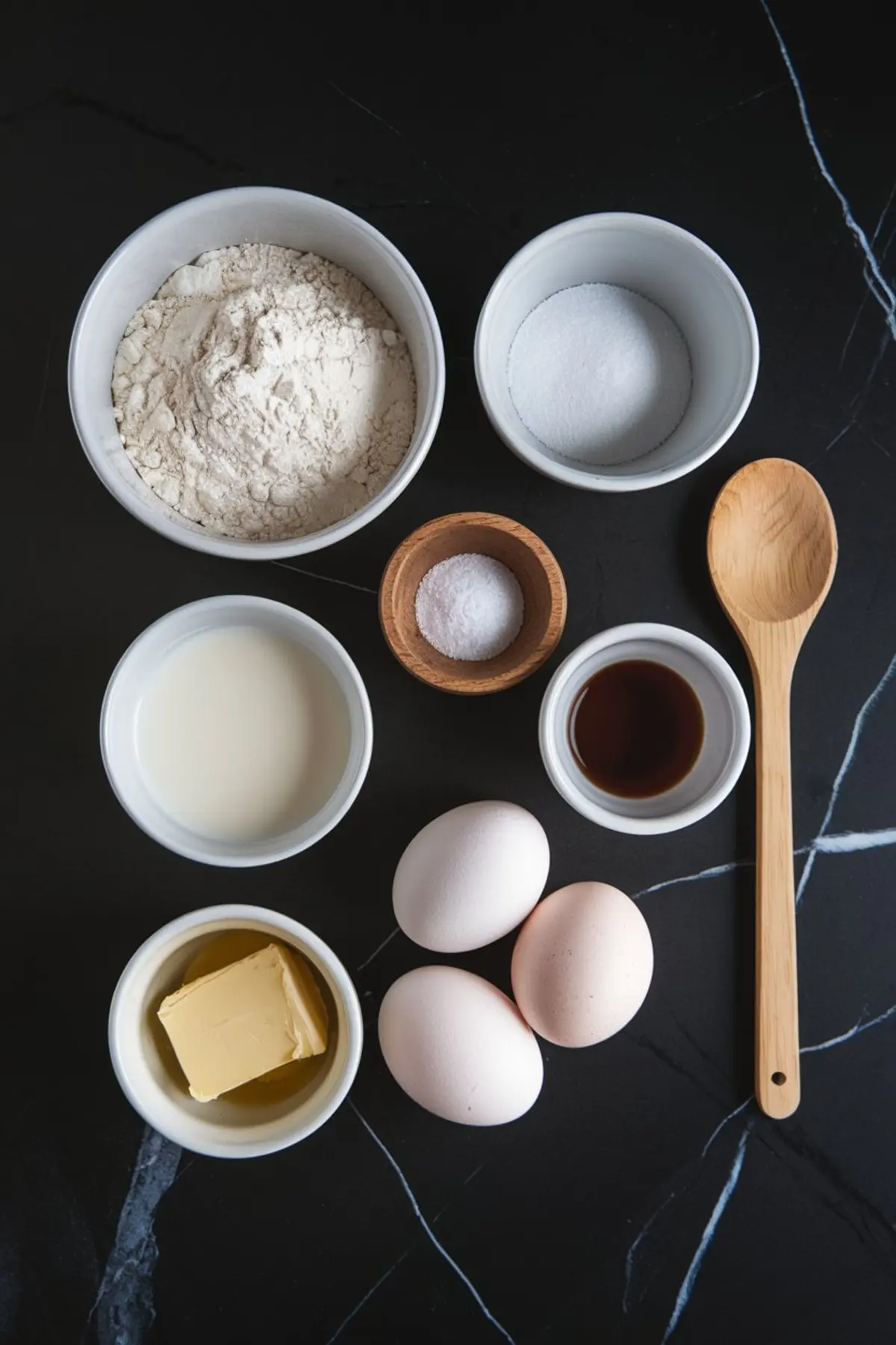 An overhead shot of neatly arranged ingredients in small bowls on a black surface, including flour, sugar, salt, milk, butter, eggs, and vanilla extract. A wooden spoon rests beside them.