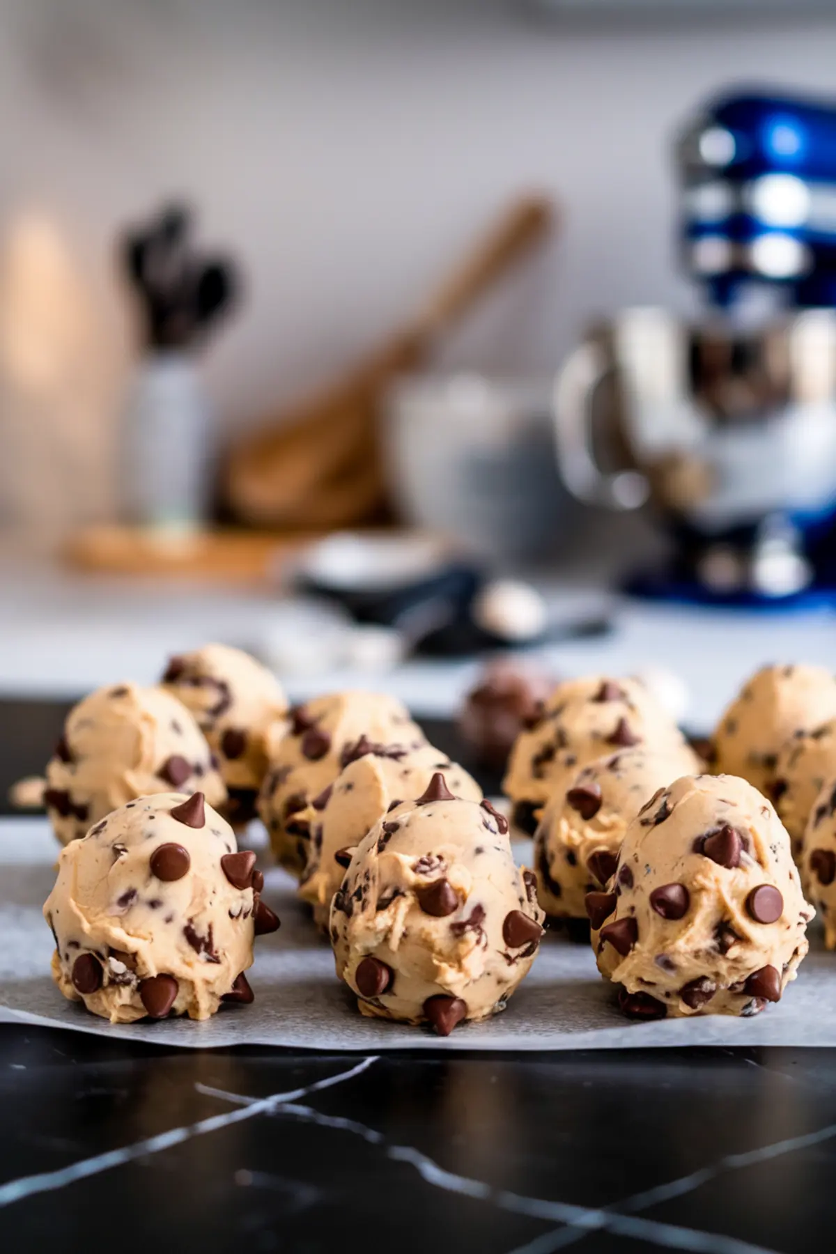 Rows of raw chocolate chip cookie dough balls resting on a parchment-lined surface. The dough is studded with chocolate chips, and the background features a blurred kitchen with a blue stand mixer and utensils.