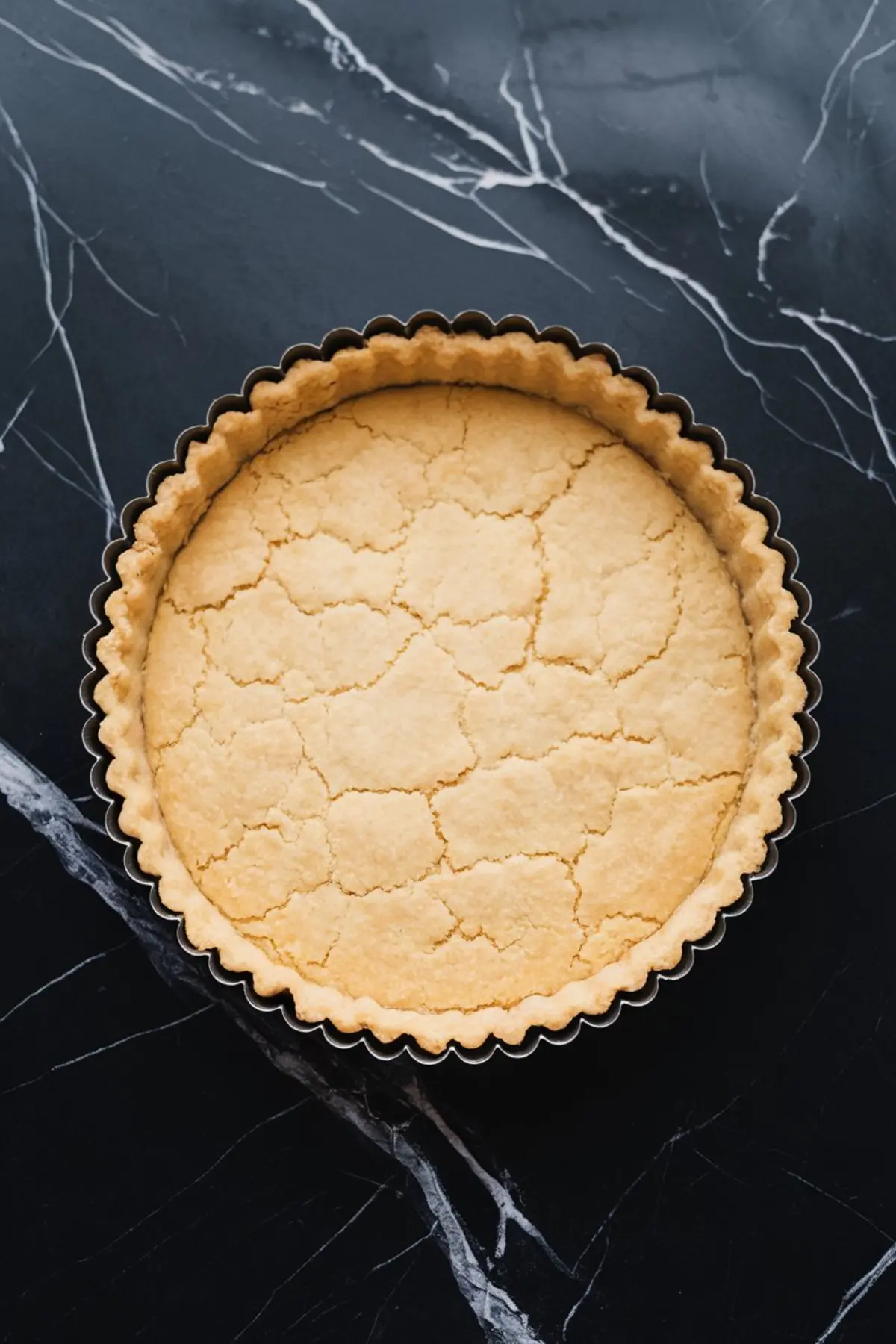 A baked tart crust with a golden, flaky appearance and slight cracking on the surface, resting in a fluted tart pan on a black marble countertop.