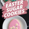 A tray of Easter sugar cookies decorated with floral designs, featuring hand-piped daisies, tiny white blossoms, and yellow flowers with green grass details. A pink egg cookie with three white daisies is placed on a white plate. Bold pink brushstroke text reads “Easter Sugar Cookies.”