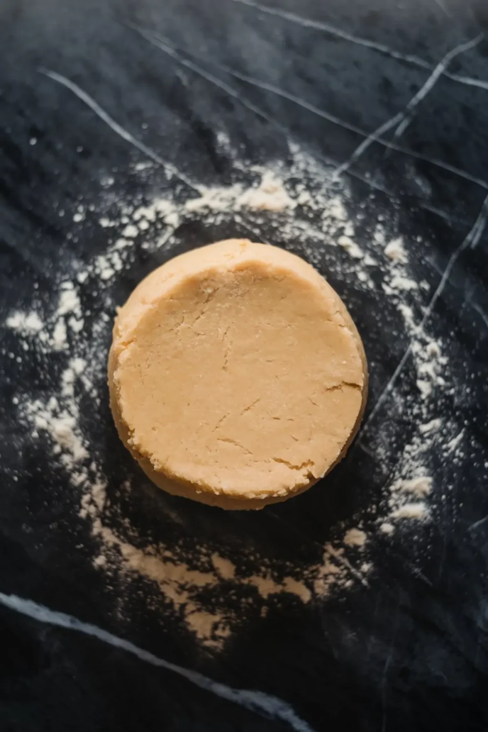 A round disc of sugar cookie dough on a dark floured surface, ready for rolling and cutting. The slightly cracked edges and light dusting of flour add a rustic homemade feel.