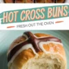 A collage of three images displays freshly baked hot cross buns with a shiny, golden-brown top, decorated with a white and dark chocolate cross pattern. The middle section features a vintage-style text banner reading “HOT CROSS BUNS FRESH OUT THE OVEN.” The bottom image focuses on a single bun with a bite taken out, placed on a ceramic plate.