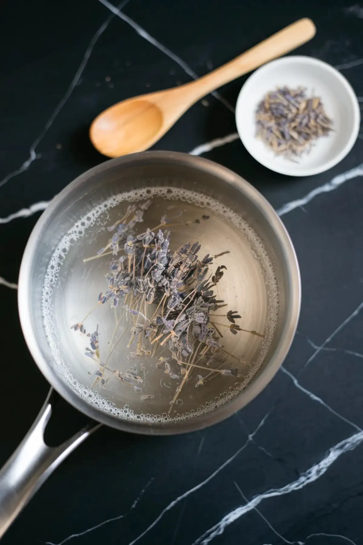 Stainless steel saucepan with dried lavender flowers simmering in water on a black marble countertop, accompanied by a wooden spoon and a small bowl of loose lavender buds.