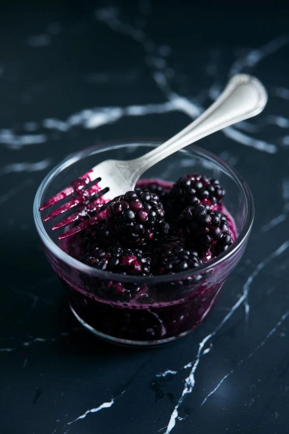 Mashed blackberries in a glass bowl with a fork, releasing rich purple juice, placed on a dark marble surface, highlighting the fruit’s natural color and texture.