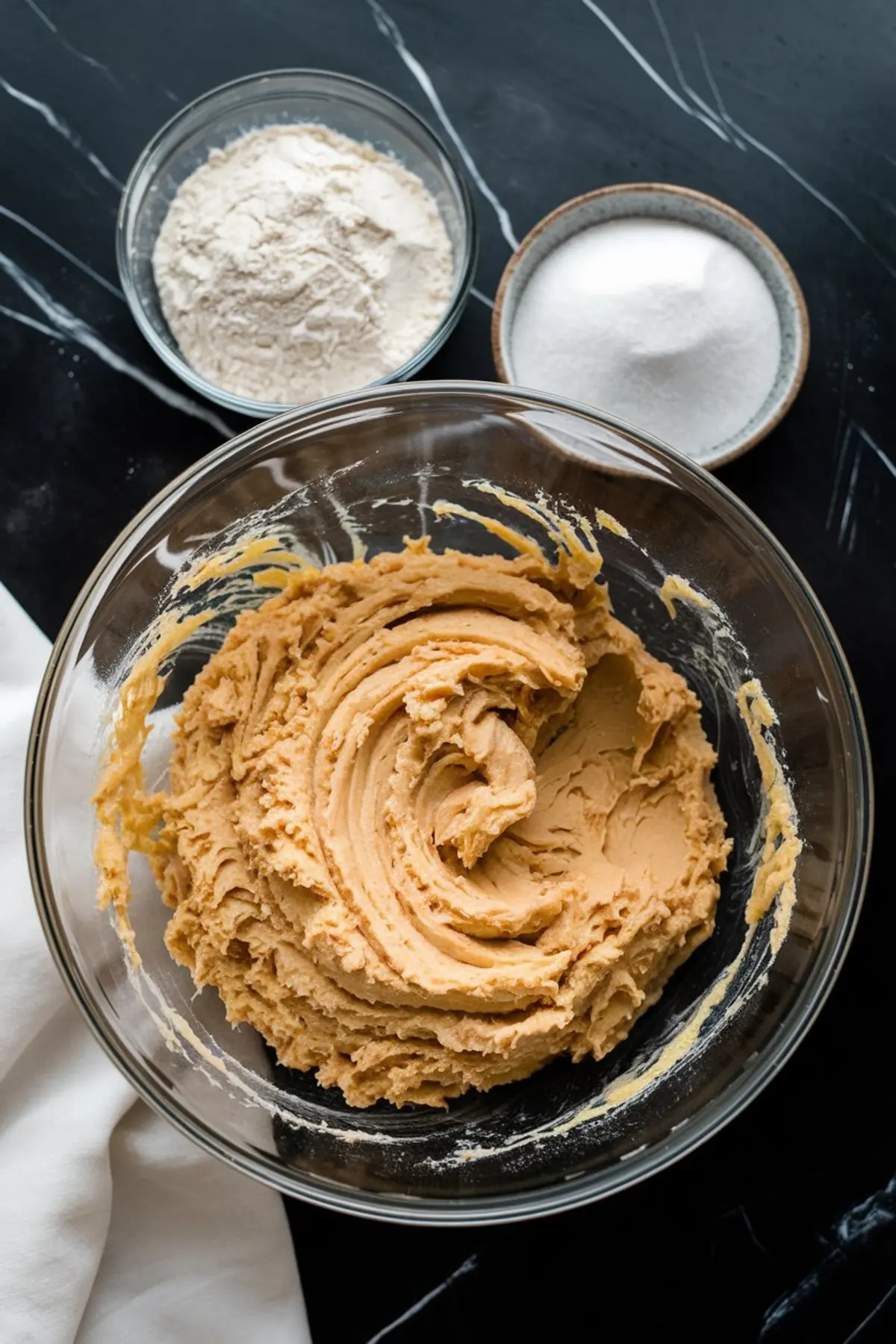 Creamed butter and sugar mixture in a glass bowl, ready for dry ingredients, with bowls of flour and sugar nearby on a dark countertop. The smooth, fluffy texture indicates the start of cookie dough preparation.