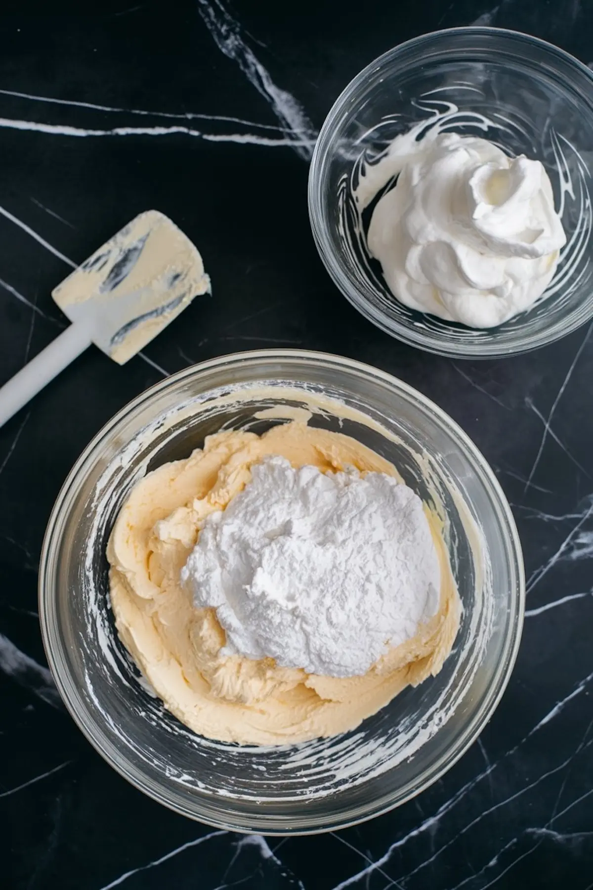 A mixing bowl filled with a light cheesecake batter topped with powdered sugar. A separate bowl holds whipped cream, ready to be folded in. A spatula coated with batter rests on the dark marble surface.