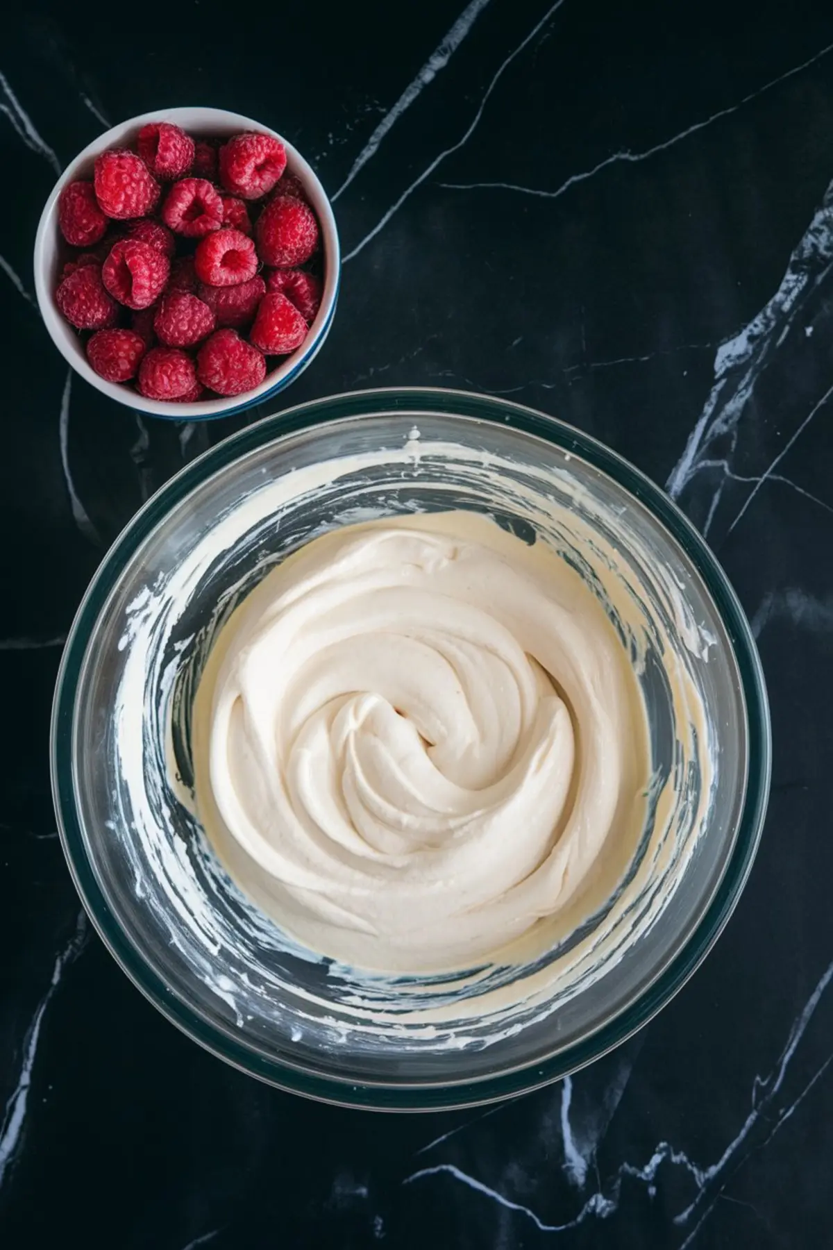 A glass mixing bowl filled with creamy white cheesecake batter on a black marble surface. Next to it, a small bowl holds fresh red raspberries. The smooth texture of the batter contrasts with the vibrant berries.