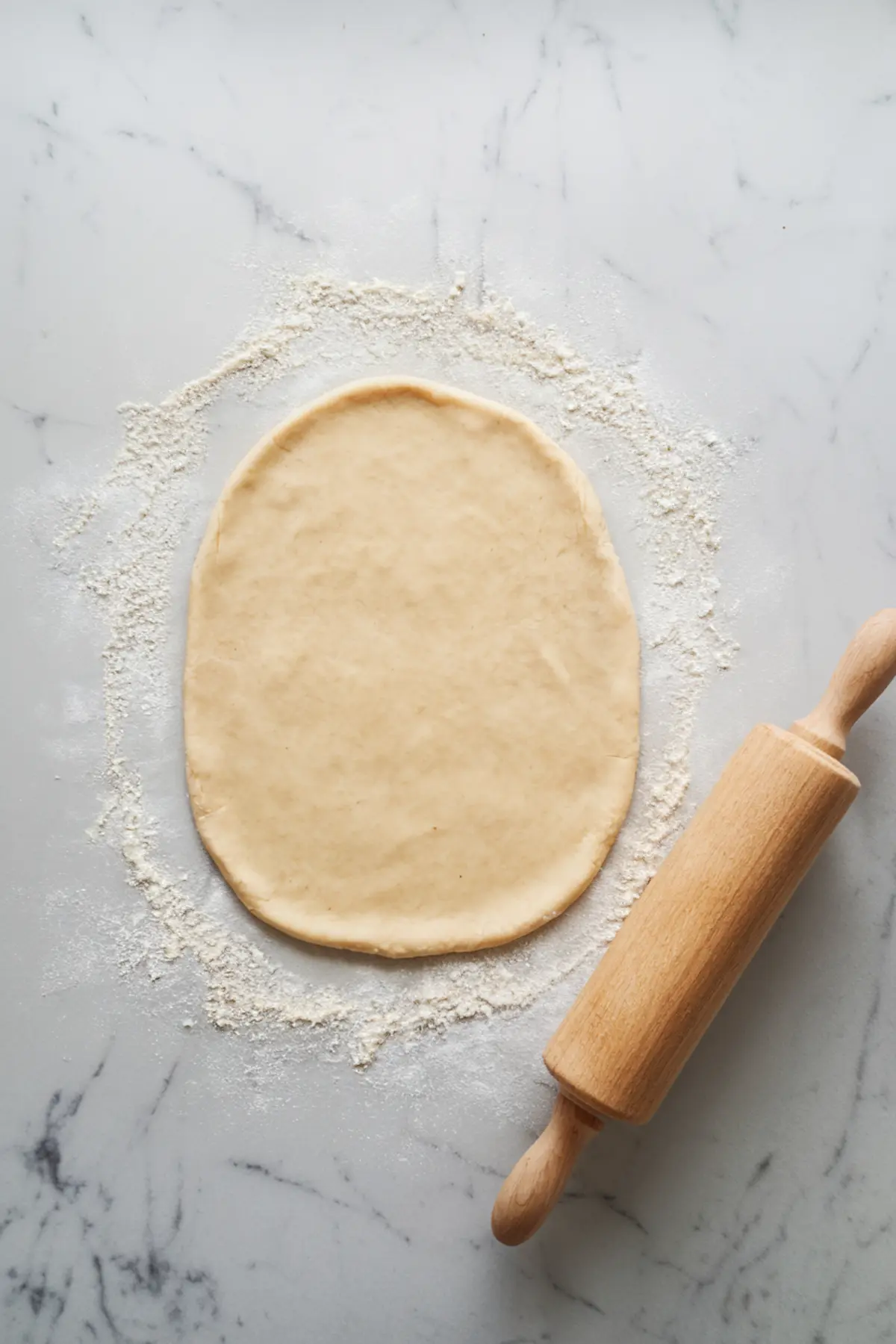 Image of rolled-out dough on a floured marble surface with a wooden rolling pin beside it, ready for shaping.