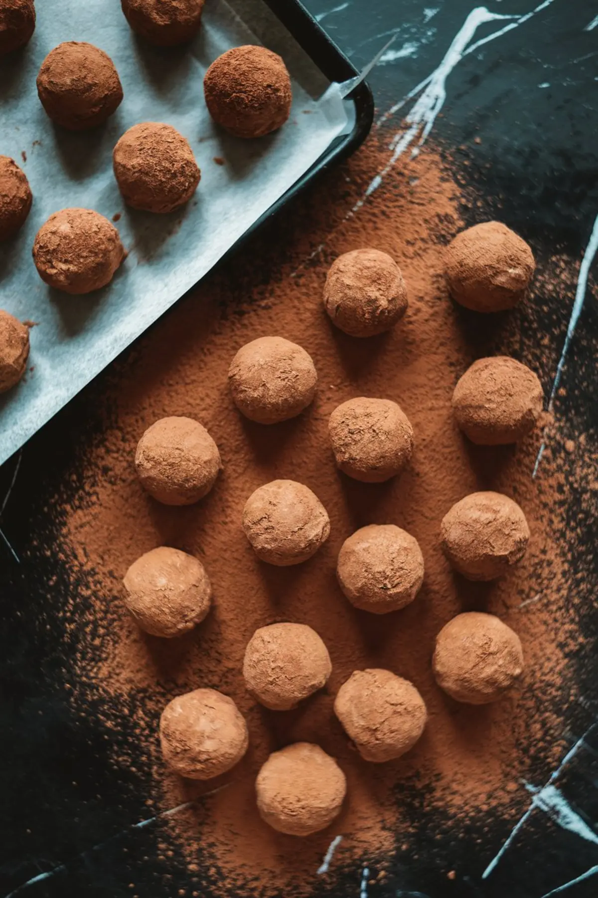 Tiramisu truffles being prepared on a dark surface. Cocoa-dusted truffles are placed on parchment paper over a baking tray, while more truffles are arranged neatly on the countertop. Cocoa powder is scattered around, enhancing the rustic presentation.