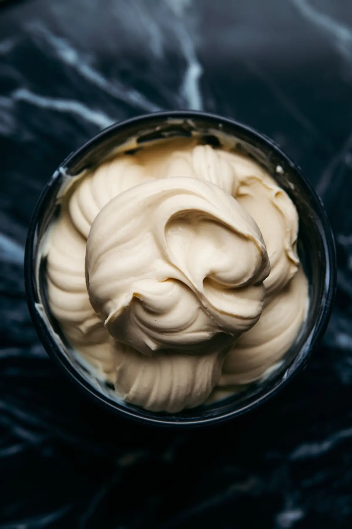 Close-up of creamy mascarpone filling in a bowl. The whipped mixture has a smooth, velvety texture with rich swirls. The dark marble background contrasts with the light-colored filling, emphasizing its creamy consistency.