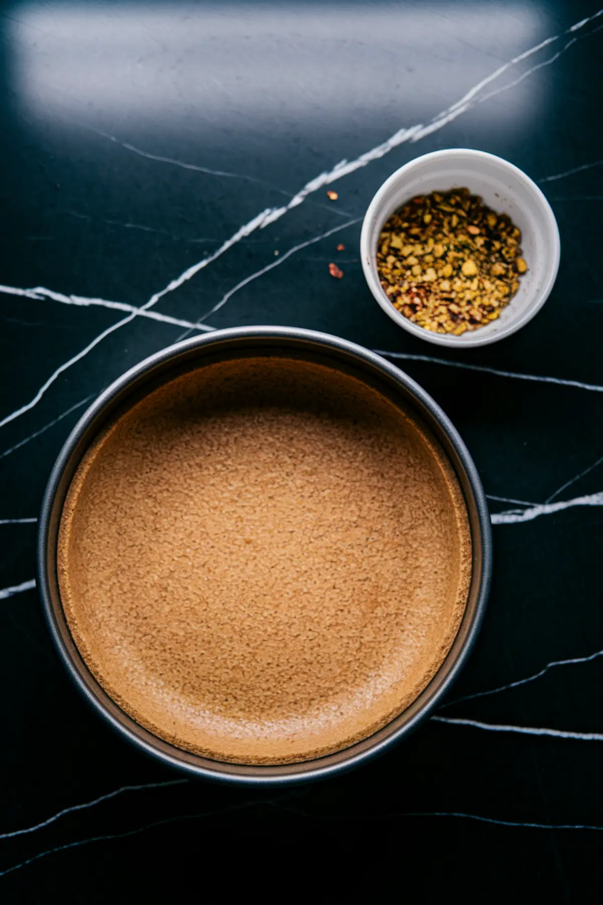 A freshly baked graham cracker crust in a round baking pan, sitting on a black marble surface. A small bowl of chopped pistachios is placed nearby, ready to be used as a topping.