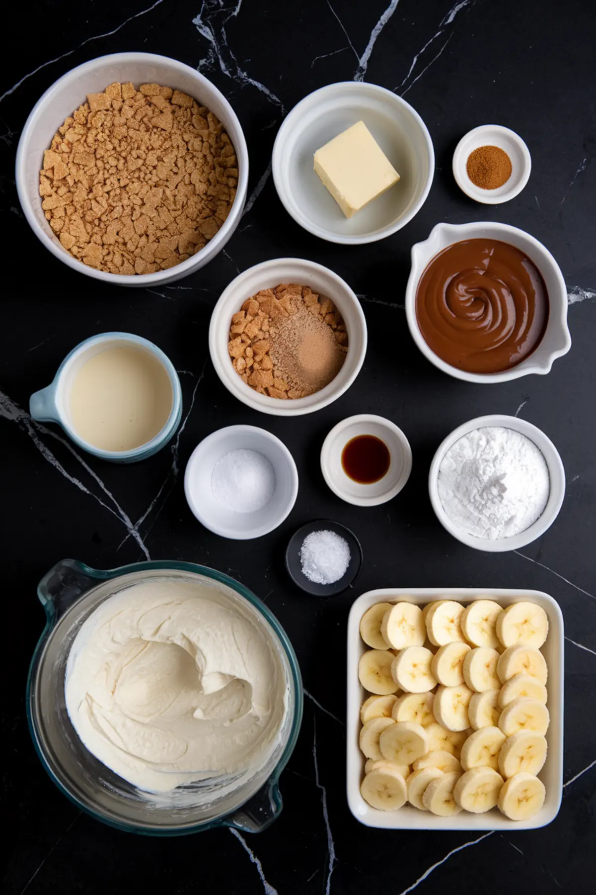 Flat lay of ingredients for banana caramel cream bars on a black marble surface. Items include sliced bananas, whipped cream, crushed graham crackers, caramel sauce, powdered sugar, vanilla extract, sweetened condensed milk, and butter.