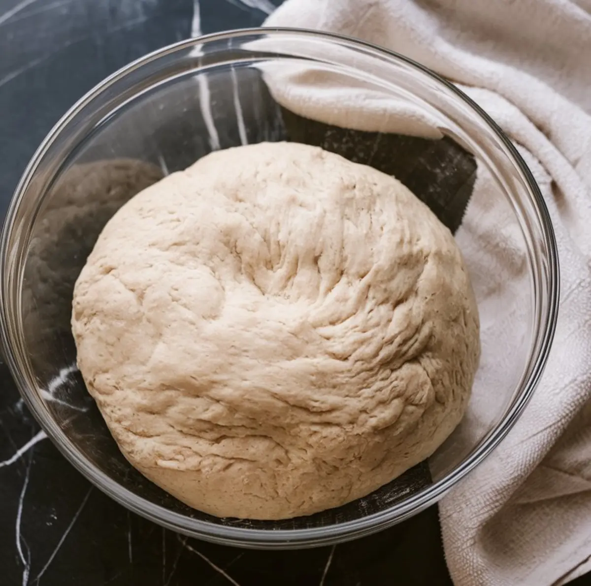 Smooth and risen banana cinnamon roll dough in a clear bowl, rested and ready for rolling, with a towel beside it on a dark marble counter.