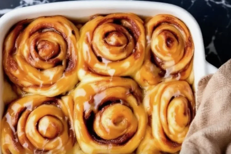 Banana cinnamon rolls baked in a white dish, swirled and golden with glossy caramelized banana glaze, set against a black marble backdrop with yellow text overlay.