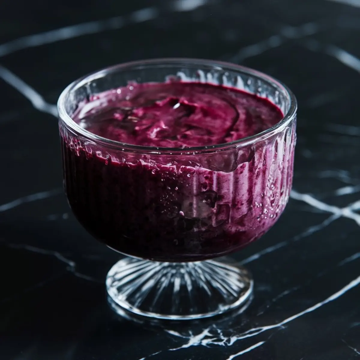 Glass dessert bowl filled with blended berry lemonade sorbet mixture, showing a glossy deep purple color and creamy texture against a black marble backdrop.