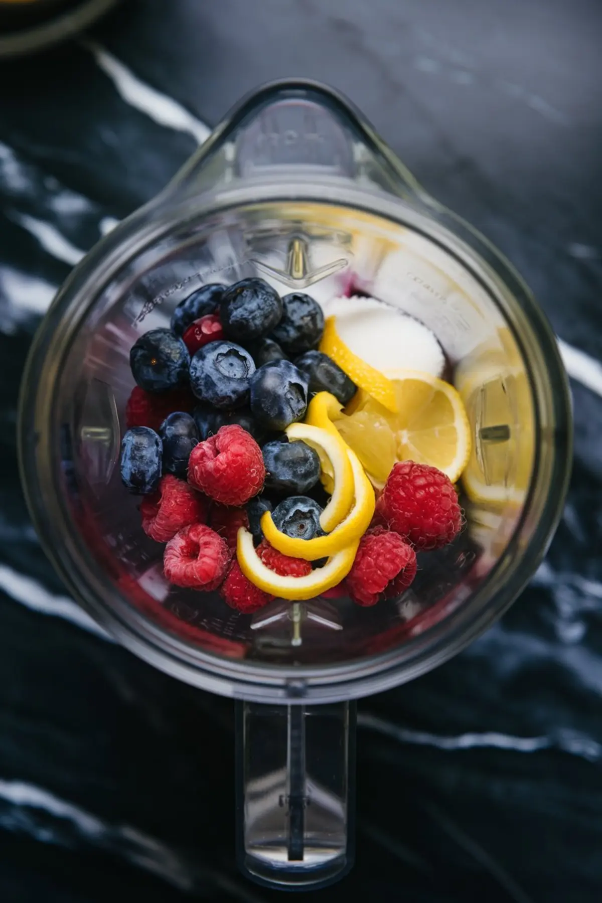 Top-down view of a blender filled with ingredients for berry lemonade sorbet, including blueberries, raspberries, lemon slices, lemon zest, and sugar, arranged for a vibrant, pre-blend preparation shot.